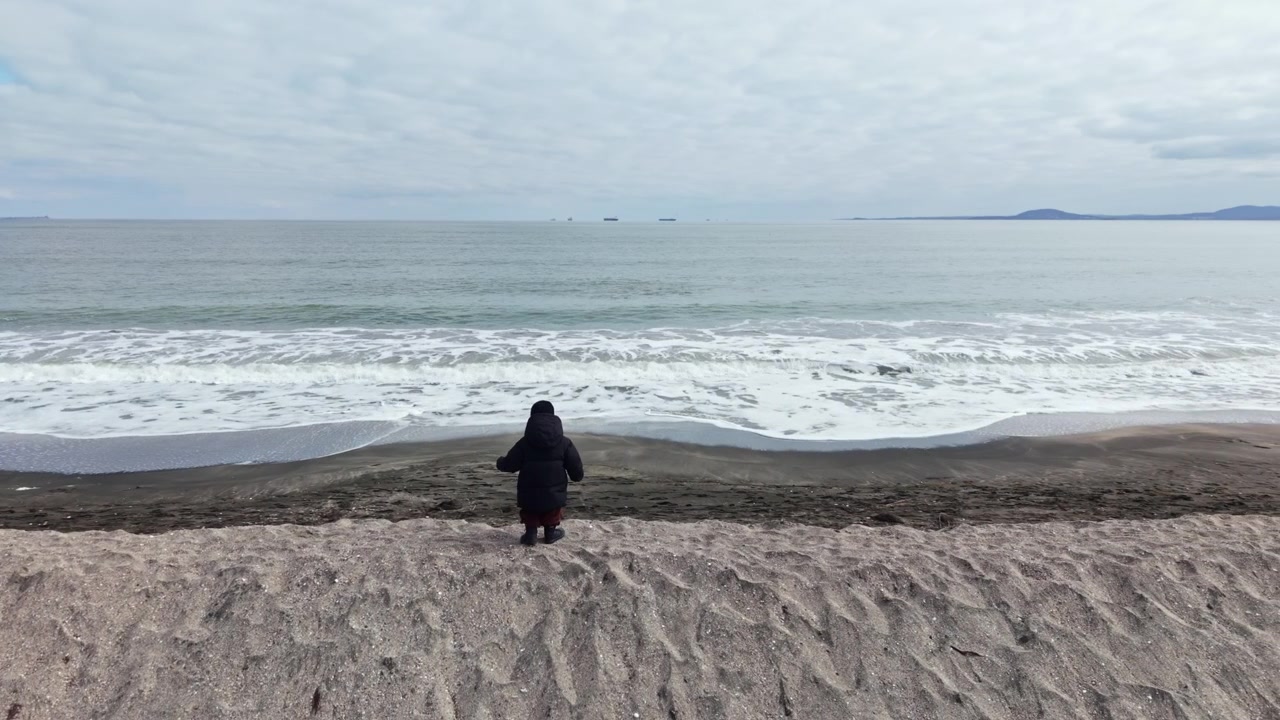 Et barn går langs en strand nær havet, mens bølgerne slår blidt mod kysten på en overskyet dag.