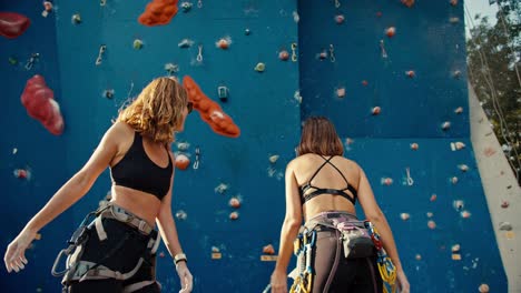 A-blonde-girl-with-curly-hair-and-a-girl-with-a-bob-hairstyle-give-each-other-a-high-five-with-hands-using-chalk-for-sports-and-begin-their-whipping-on-a-blue-climbing-wall-in-the-summer