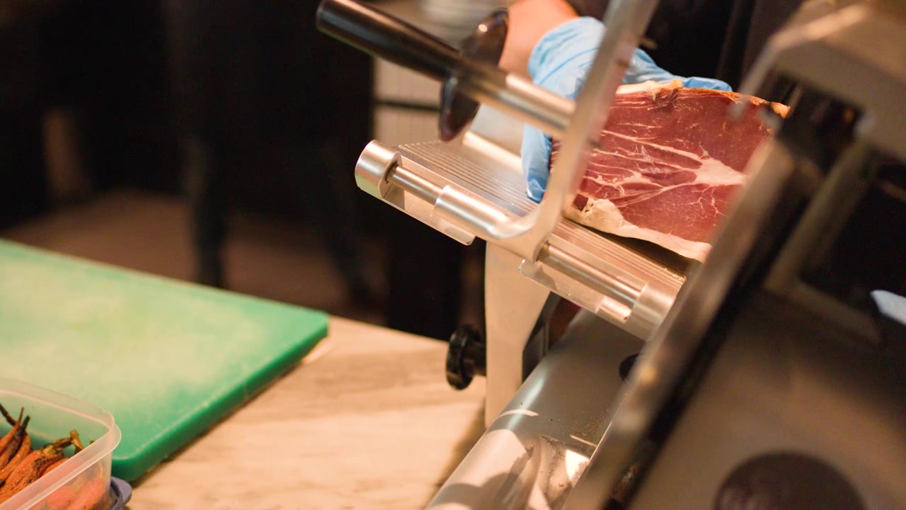 Close-up of chef using electric slicer on cured ham in a professional restaurant kitchen.
