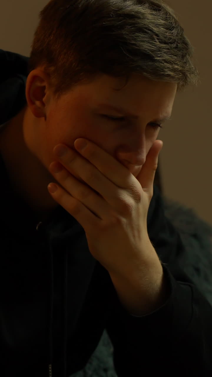 Jovem cobre a boca em um quarto escuro, demonstrando profunda tensão emocional e luta interna, capturado em um close-up cinematográfico