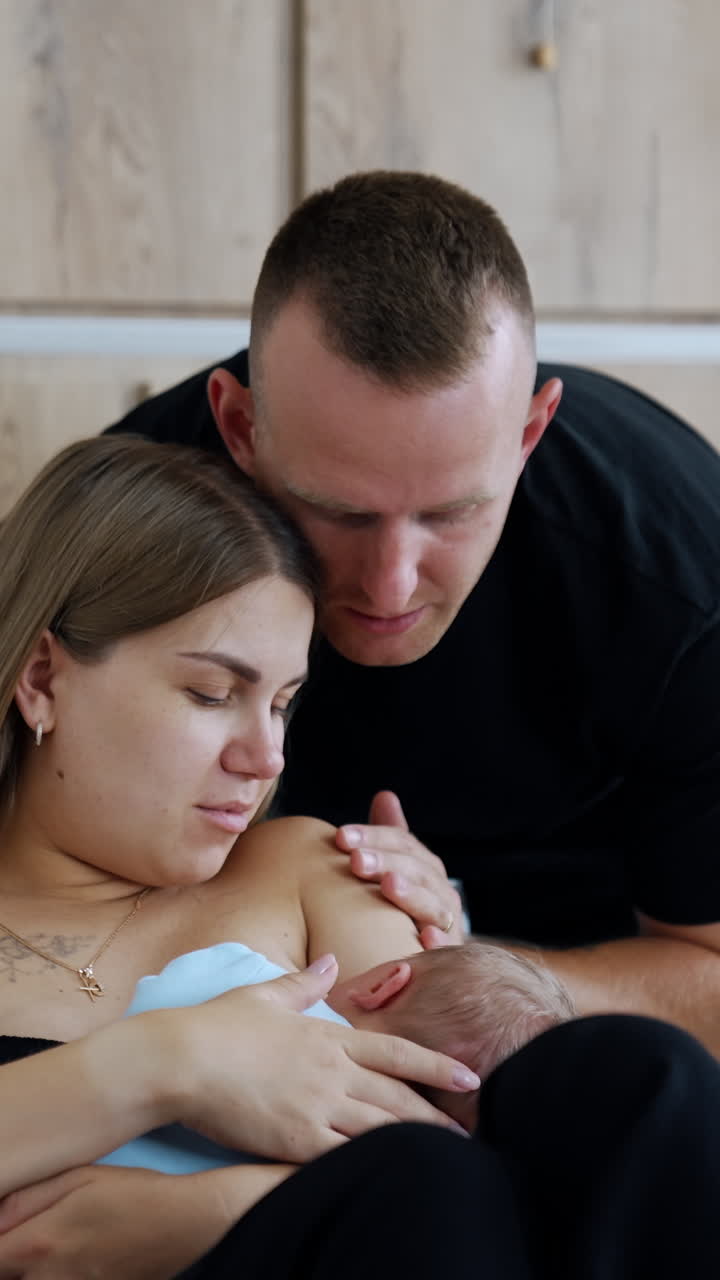 New-made mother breastfeeding her newborn child. Happy father stands behind bending over the wife closer to kid. Vertical video