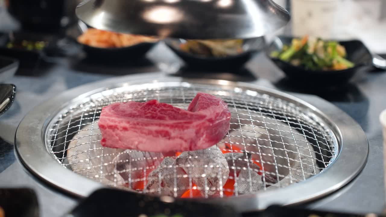 Close up of luxurious raw Hanu beef steak and king oyster mushroom grilling slowly over hot charcoal in an authentic Korean barbecue restaurant setting surrounded by side dishes