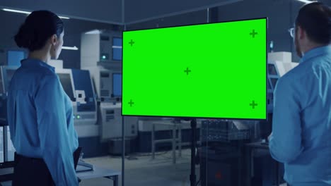 kantoorvergadering: zelfverzekerde vrouwelijke ingenieur praat met projectmanager, kijkt naar een interactieve digitale whiteboard-tv met een groen scherm en een chroma-sleuteldisplay. moderne fabriek met machines