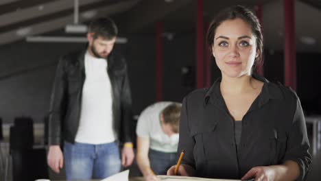 Portrait-of-a-fashionable-young-professional-woman-holding-folders-with-papers-and-taking-notes-in-a-stylish-contemporary-office.-Looking-at-the-camera.-Office-workers-on-the-background.-Shot-in-4k