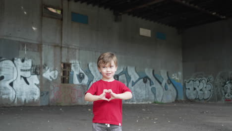 a happy young little boy is having fun and making heart symbols with hands in front of camera in a red t-shirt. young boy standing in front of a graffiti wall at a abandoned place.