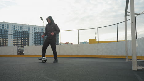 athlete practicing soccer drills on sport arena, skillfully controlling ball under soft daylight, background includes urban residential buildings, lampposts, goalpost, and mesh fencing