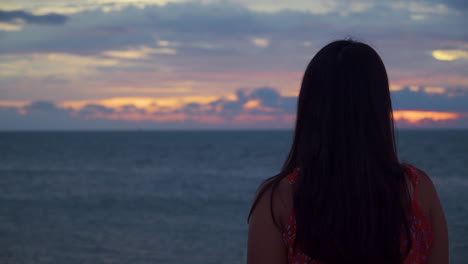 rear view of pretty woman in sundress watching amazing colorful dramatic sunset over endless sea