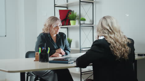 client adjusts forward paying attention to what lady is showing her during professional meeting in office space includes organized shelf with green plants pen holder and digital tablet on table