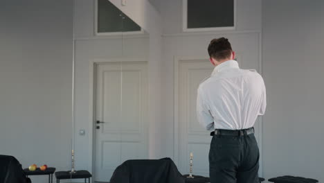 rear view of man dressing up in crisp white shirt and black belt in front of large mirror in bright loft studio interior, reflecting double doors, capturing collar adjustment and poised movement