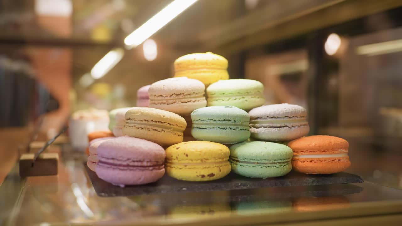 close up colorful macarons stacked neatly in dessert display case under warm lights with faint silhouette of person walking by reflected in glass surface
