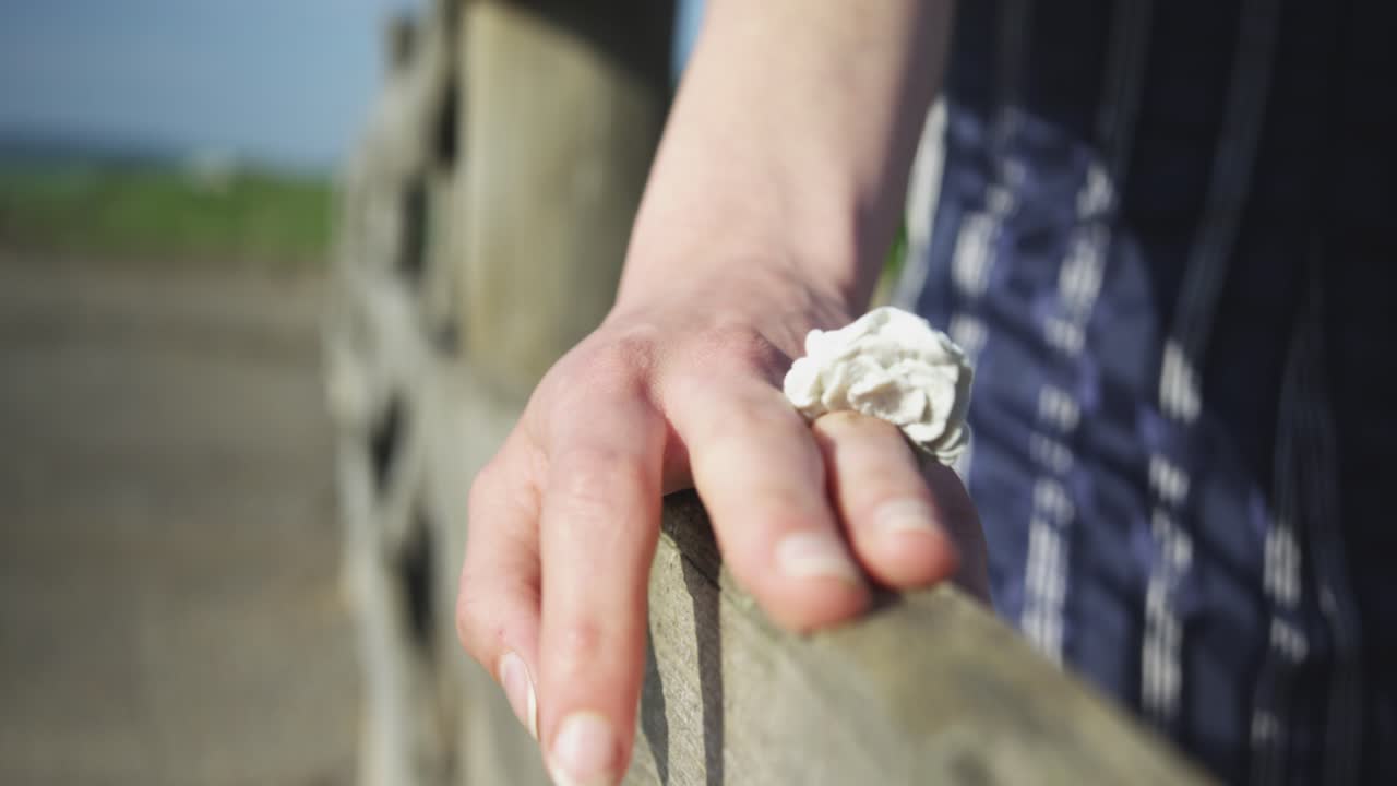 A close-up, handheld shot follows a woman's hand, adorned with a large white ring, as it gently slides along the top of a rustic wooden fence in the countryside on a sunny day.