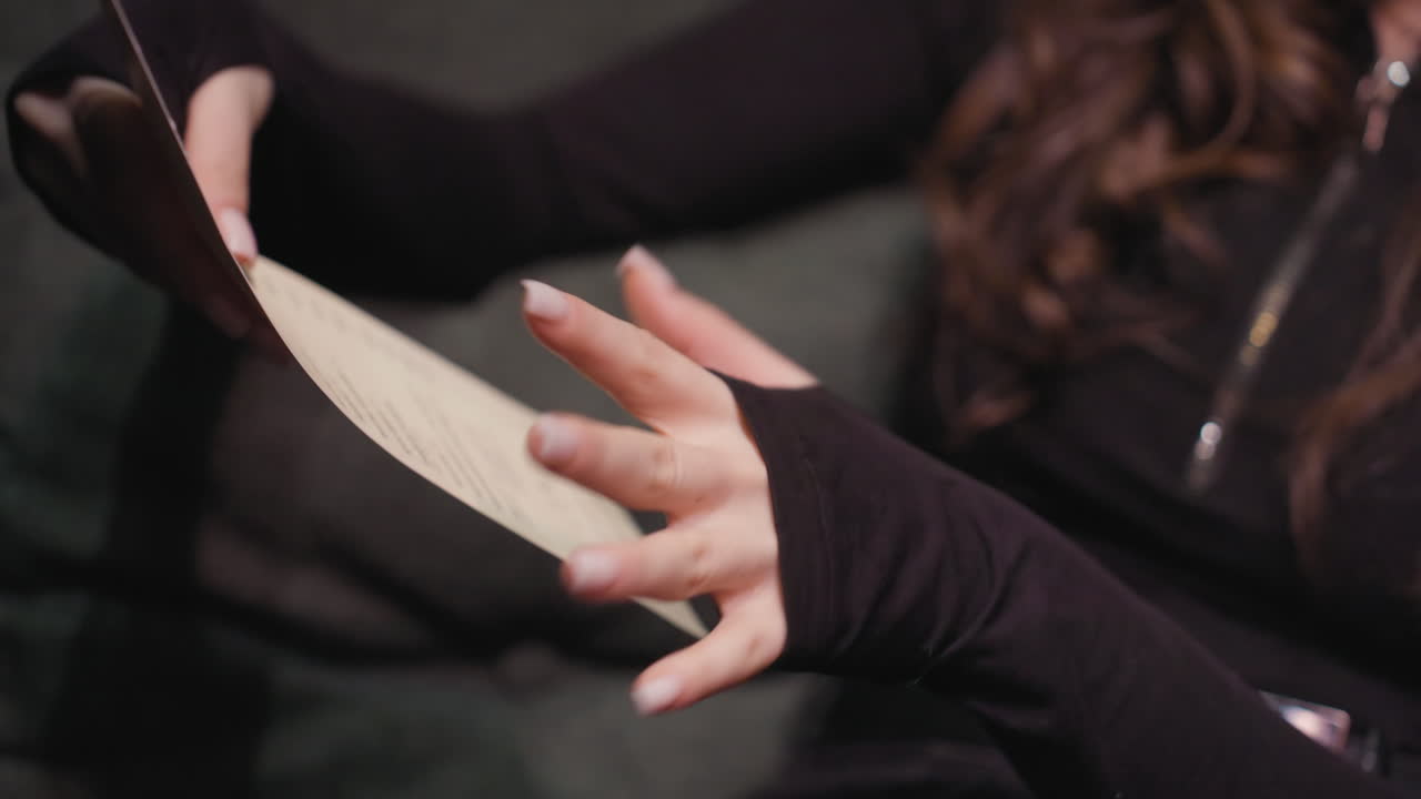 Close up view of woman in black outfit holding menu and pointing at selected item with her finger. Scene captures intent and engagement in cozy setting with soft light and dark green cushion