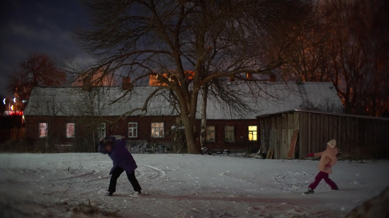 Child running through snow in front of old brick building at night. Winter scene in Daugavpils with bare trees, snow covered yard, and warm light from windows of traditional regional house.