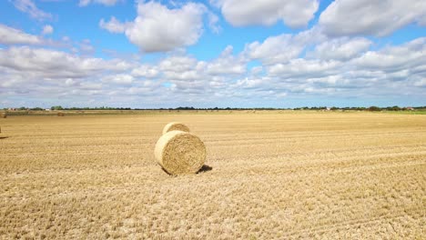 Hoch-über-Der-Landschaft-Wirbelt-Eine-Reihe-Von-Windkraftanlagen-Elegant-Auf-Dem-Frisch-Abgeernteten-Feld-Eines-Bauern-Aus-Lincolnshire,-Wobei-Goldene-Heuballen-Die-Aussicht-Verschönern