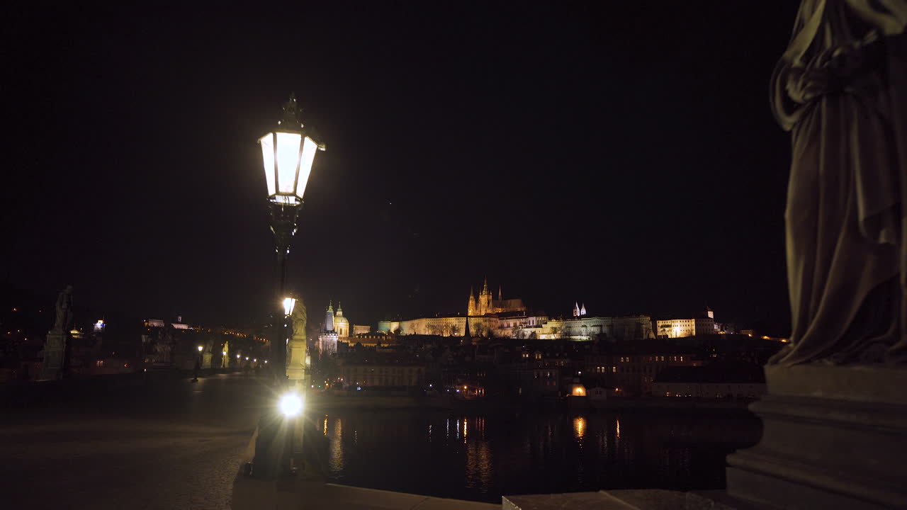 una cruz de piedra ornamentada con jesús crucificado en el puente de charles en praga por la noche durante un encierro covid-19, estatuas y linternas a ambos lados, castillo de praga iluminado en el fondo, tiro de 4k