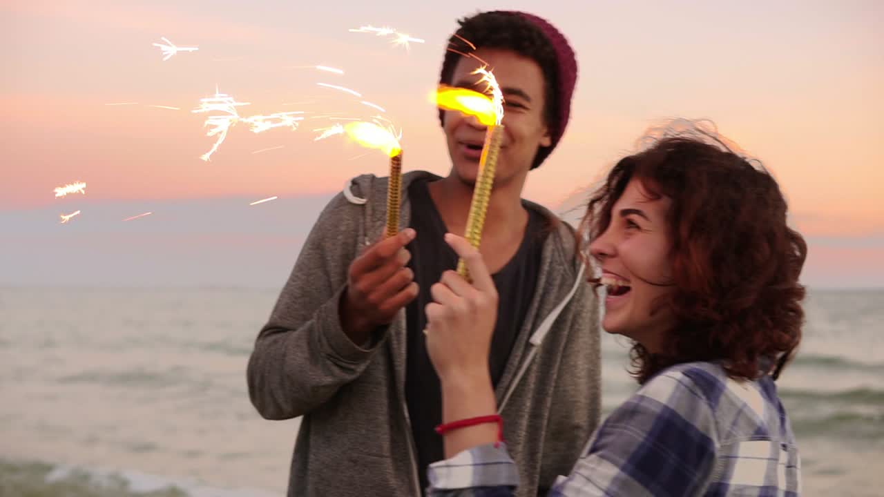 alegre pareja multiétnica sonriente sosteniendo velas brillantes encendidas de pie junto al mar durante el atardecer