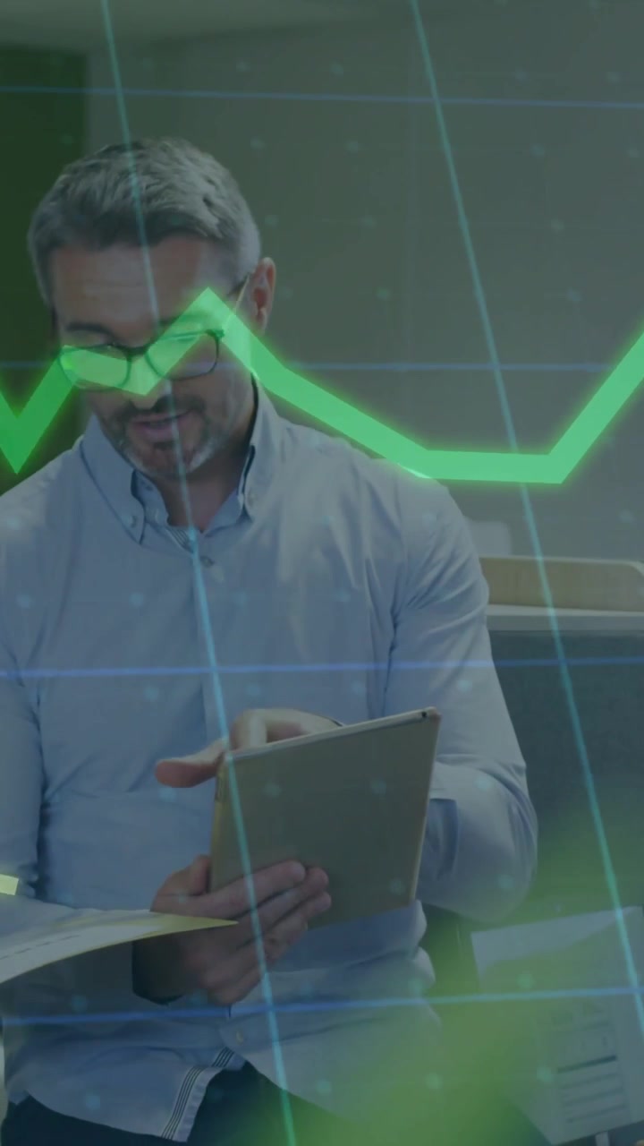 Uomo con occhiali e camicia, seduto in ufficio, che tiene un tablet e una cartella e studia grafici verdi e blu.