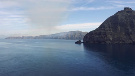 Majestuosos-Acantilados-Marinos-Vigilan-Las-Oscuras-Aguas-Azules-De-Santorini,-Grecia,-Con-Casas-Blancas-En-Los-Acantilados-A-Lo-Lejos