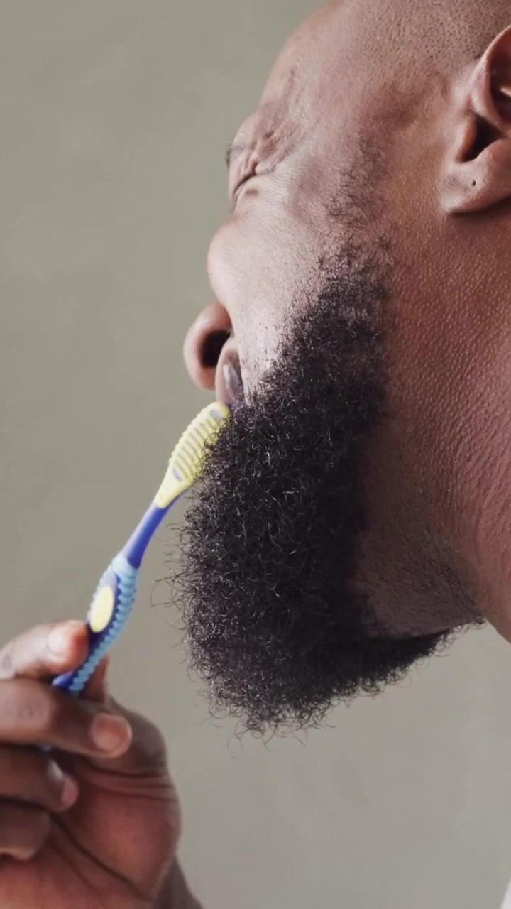 Vertical video: Grimacing man brushing teeth with toothbrush at bath sink, scrubbing beard in tank
