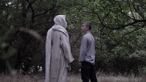 Teen boy stands in forest looking at robed man