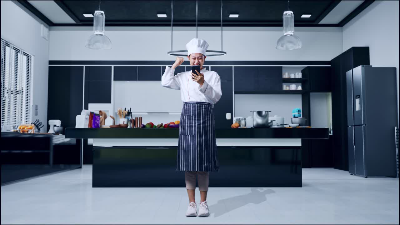 Figura intera di una chef asiatica che guarda lo smartphone, poi urla gol e festeggia nella cucina di casa