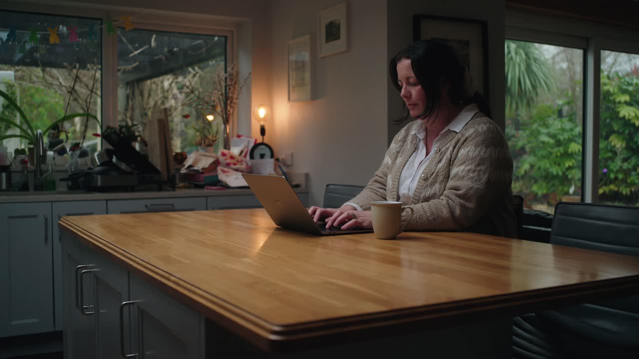 A middle-aged woman works on her laptop in a cozy kitchen, using an AI chatbot for family budgeting. Warm lighting, homely atmosphere, and focused expression.