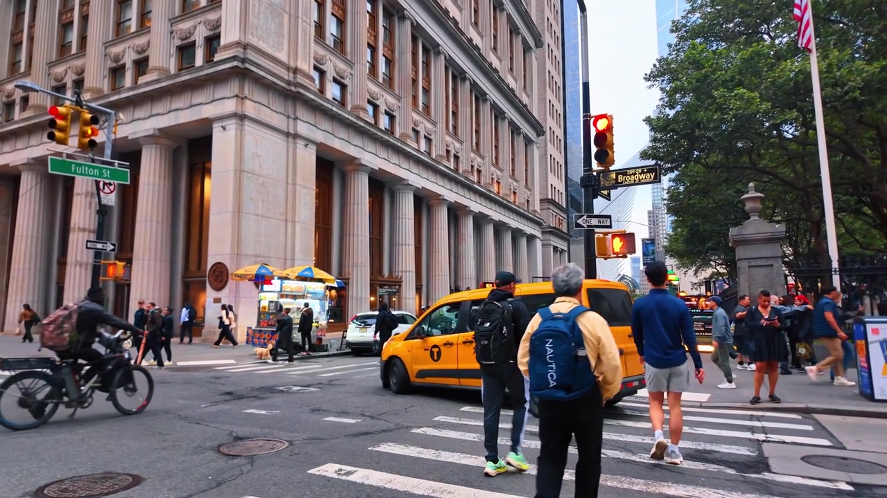 New York, USA, 5 June 2026 : Pedestrians crossing Broadway at Fulton Street, lower Manhattan. Ground view of crosswalk, yellow taxi, food cart and neoclassical building facade under overcast sky.