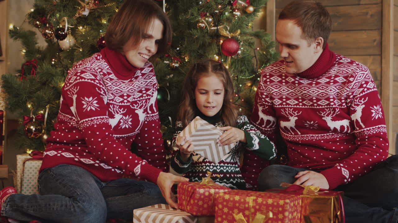 niña feliz desenvolviendo el regalo mientras se sienta con su mamá y su papá frente al árbol de navidad en casa