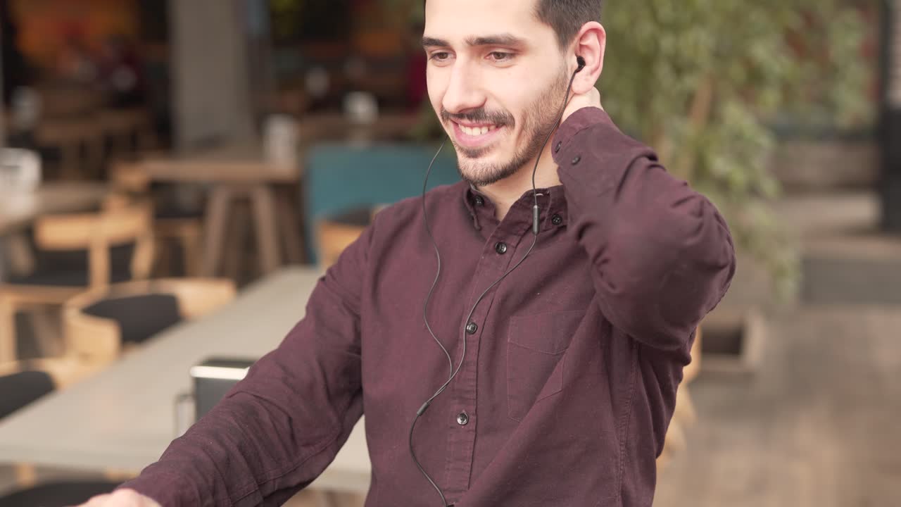 un joven con barba y bigotes está sentado en un café con auriculares, escribiendo en una laptop frente a él y sonriendo para sí mismo