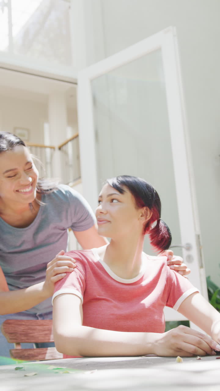 Vertical video of biracial sisters sitting at table and embracing, in slow motion
