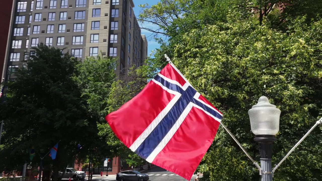 Norjan lippu liehuu koristeellisesta lyhtypylväästä historiallisen tiilirakennuksen ja modernin kerrostalon vieressä puiden reunustamalla kaupunkikadulla, muiden kansallislippujen ja pysäköityjen autojen kanssa aurinkoisen taivaan alla.