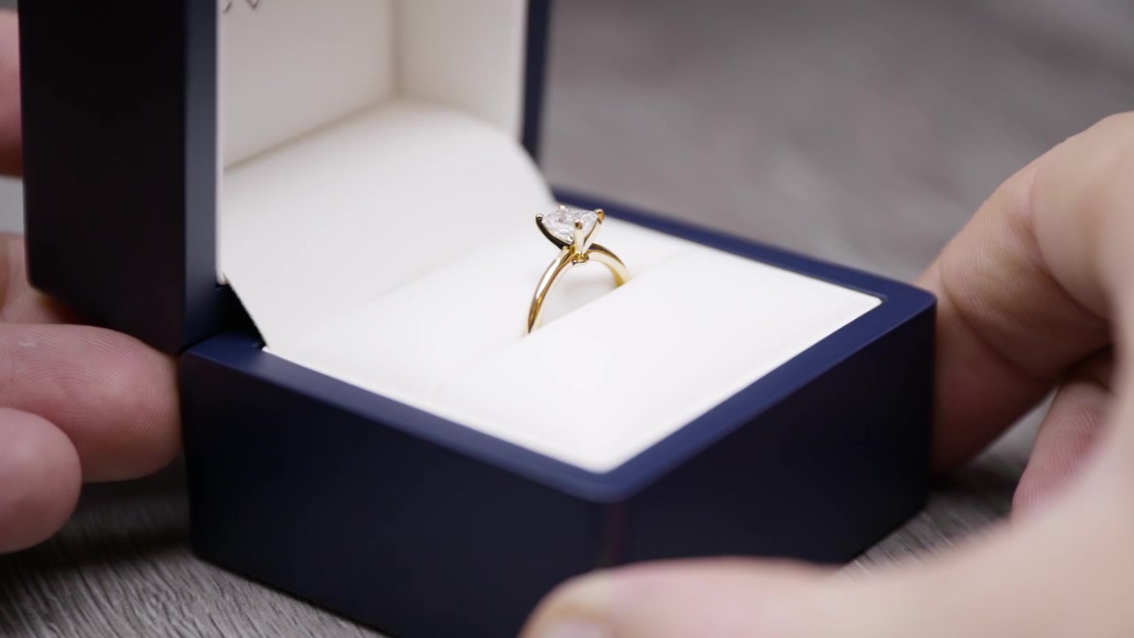 Ruce otevírající krabičku se zásnubním prstenem. Svatební prsten v otevírané krabičce. Odhalený zásnubní prsten, žádost o ruku. Diamant smaragdového brusu, žluté zlato s ostrou hranou.