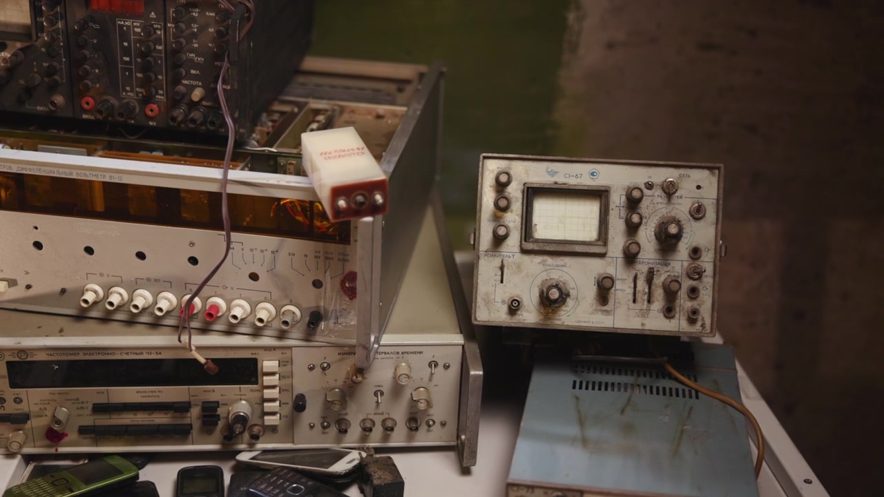 Close-up view of stacked vintage electronic test instruments beside scattered old mobile phones on cluttered workshop bench, showing cables, switches, screens, metal wear, and retro repair atmosphere