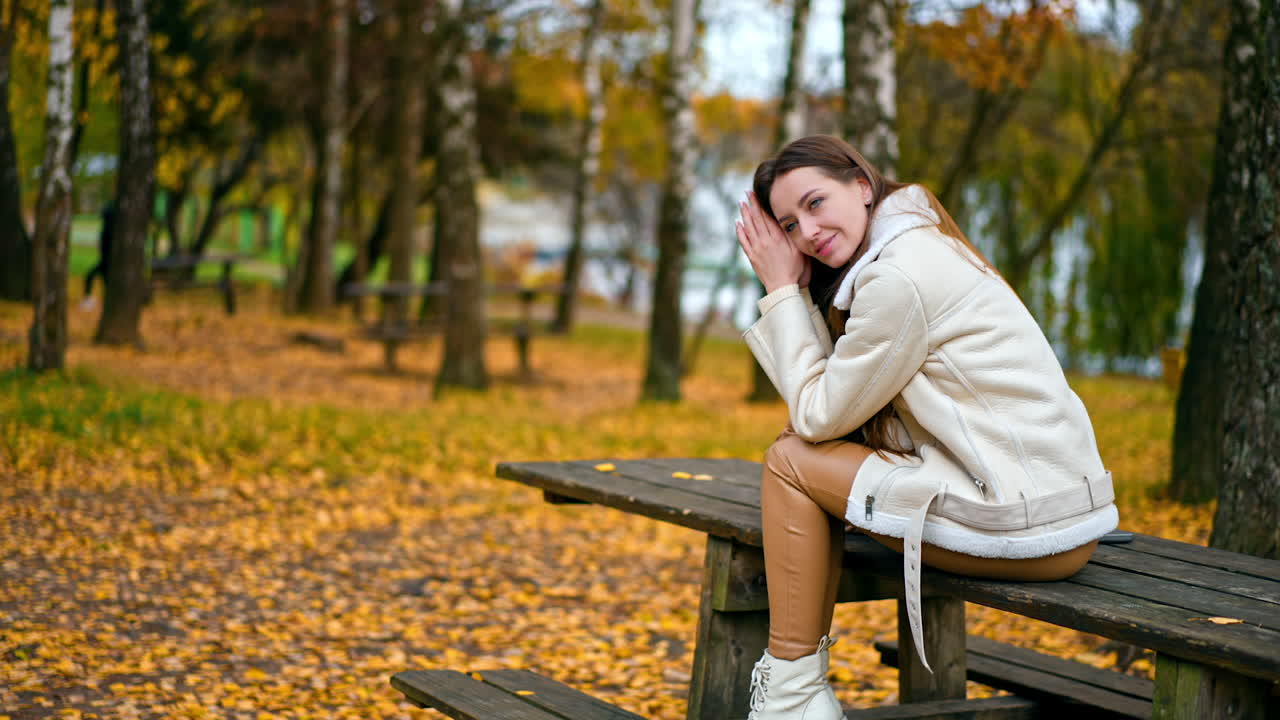 Relaxed calm dark-haired lady sits on the table thinking of something. Caucasian girl enjoying beautiful autumn day in the park among birch trees.