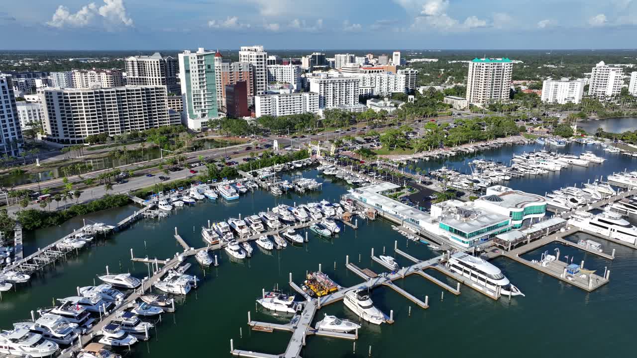 Luxurious Marina bay port of Sarasota, Florida. Aerial backwards shot. Parking and anchored boats and yachts in expensive waterfront port. Luxury hotels and apartment towers in background.