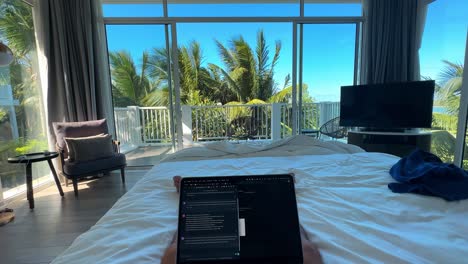 pov of wealthy individual with laptop on king-sized bed in luxurious bedroom with open balcony and palm trees