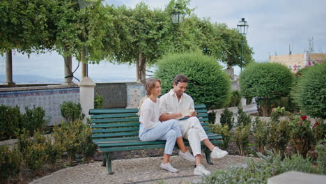 smiling newlyweds reading book at greenery square bench. lovers talking together