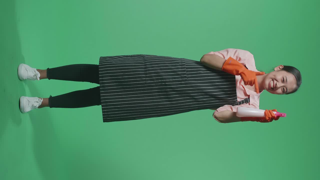 초록색 스크린 스튜디오에 서 있는 동안 스프레이를 가리키는 앞옷을 입은 미소 짓는 아시아 여성 집주인이 가득 차 있습니다.