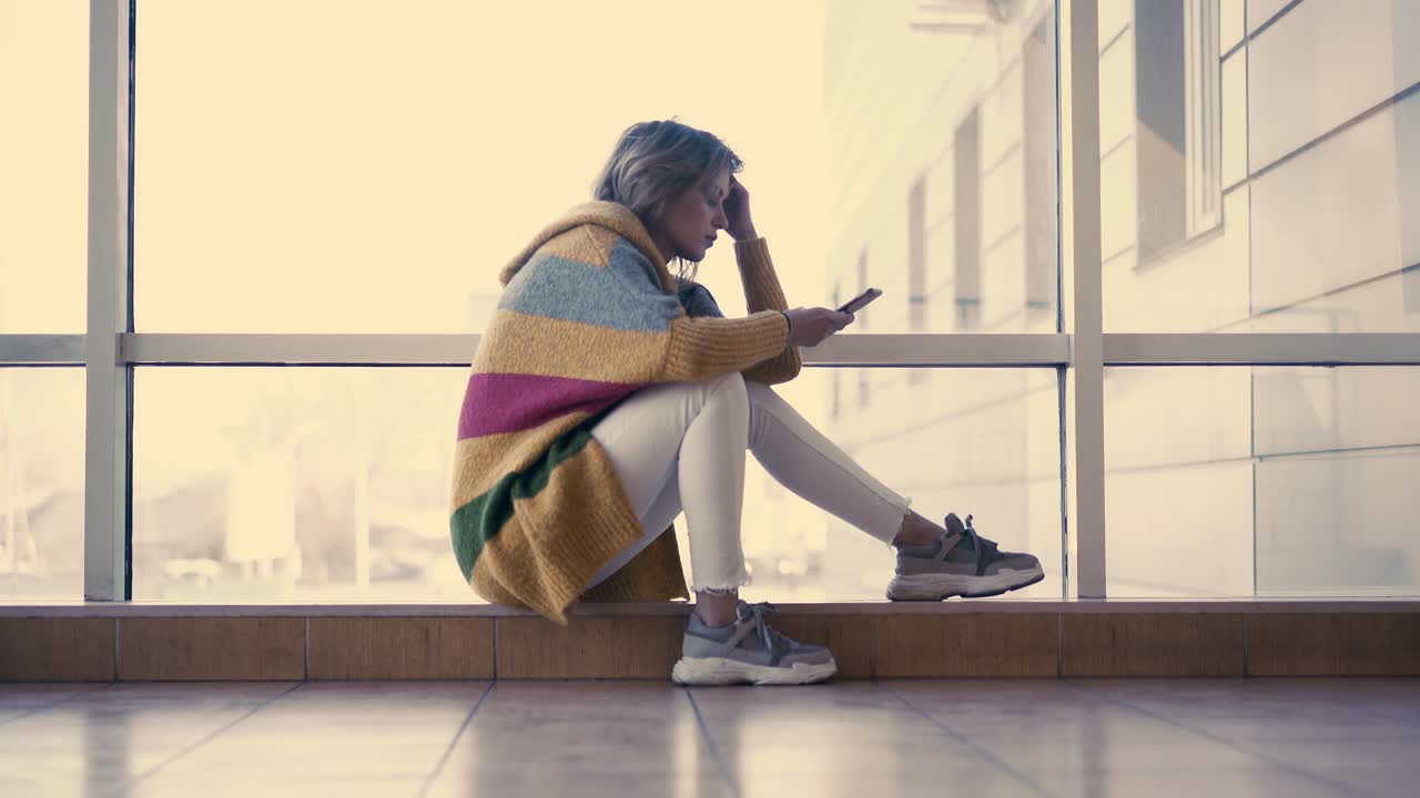 una niña linda se sienta en una ventana con un teléfono inteligente en la mano mirando pensativo al teléfono