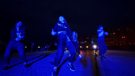 teenage boy and girls dancing hip hop outdoors at night. low angle view. people at backdrop dancing near the car with lights on.