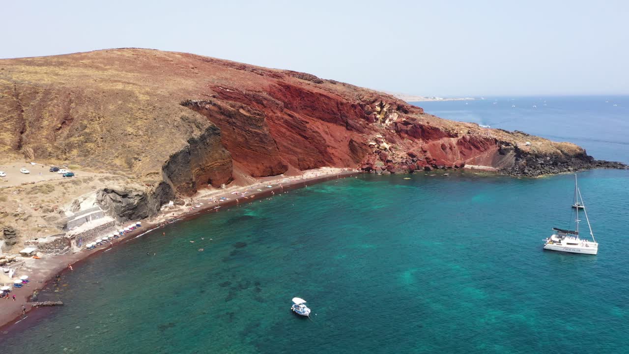 drone aéreo que revela playa roja con agua turquesa, botes, montañas y arena de color rojo en santorini, grecia