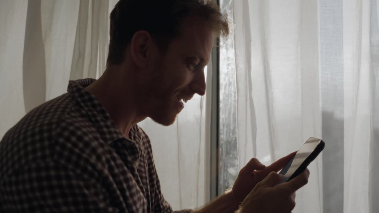 Excited and happy man typing on smart phone keyboard and texting with big smile. Cheerful and happy young person standing by window creating silhouette writing fast holding mobile phone and smiles