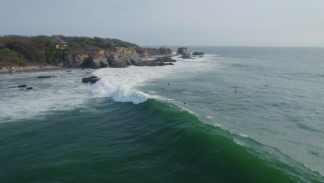 Surfers-surfing-next-to-rocks,-over-Rough-sea-with-waves,-Aerial-shot-from-the-side