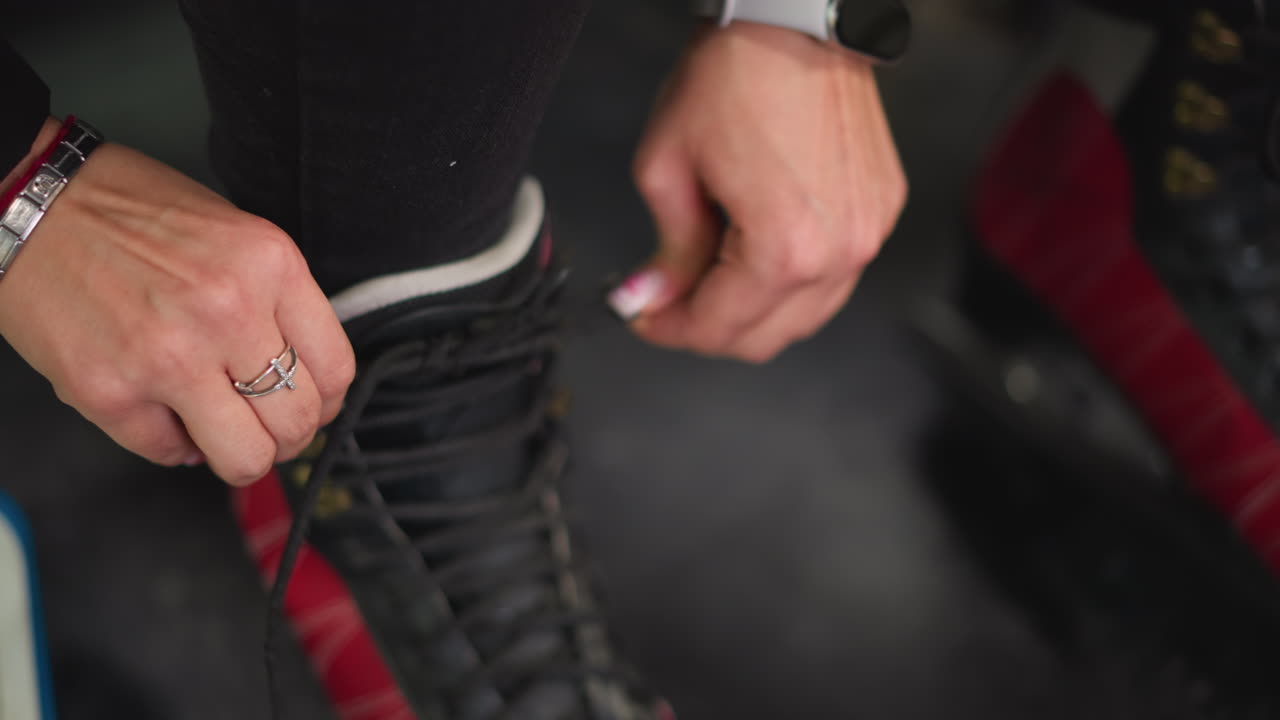 Lady putting on red black ice skates tightening laces with detailed view of hands and fingers pulling strings preparing for skating activity showing focus determination and readiness for winter sport indoors