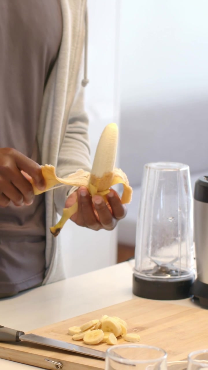 Video verticale: Uomo con felpa grigia con cerniera che prende una banana, la sbuccia sul bancone per il frullatore.
