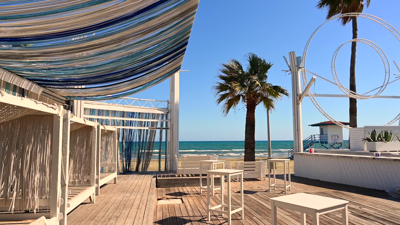 View of a beach lounge in Larnaca, Cyprus, with white wooden furniture, flowing canopy fabric, a palm tree, and the Mediterranean Sea in the background