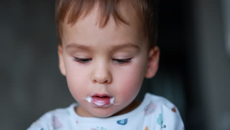 charming baby boy with dairy around his mouth. face of a kid eating yogurt from spoon. close up.