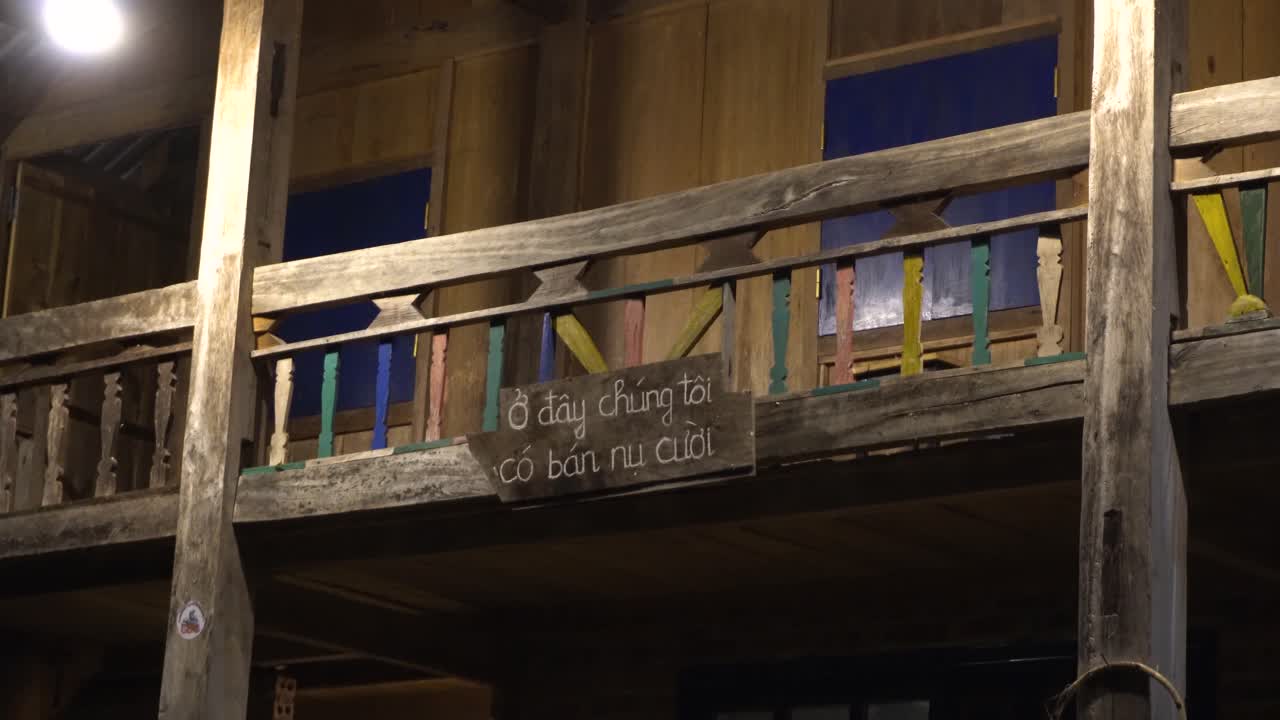 A traditional wooden balcony featuring colorful railings, illuminated warmly at night, evoking rustic charm and handmade craftsmanship. A small sign written in Vietnamese says "Here we sell smiles"