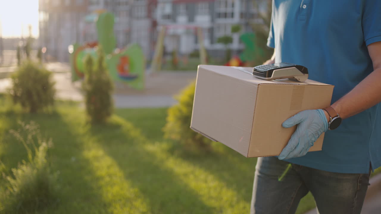 un repartidor masculino con guantes trae una caja y da la terminal para pagar la compra. llevar una caja con una terminal de pago de tarjeta de crédito nfc o teléfono móvil. pago sin contacto
