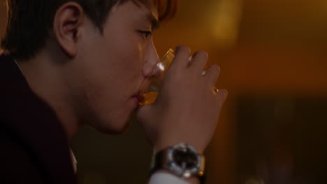 a young man drinking alone in a dimly lit room, holding a glass of whiskey, looking sad